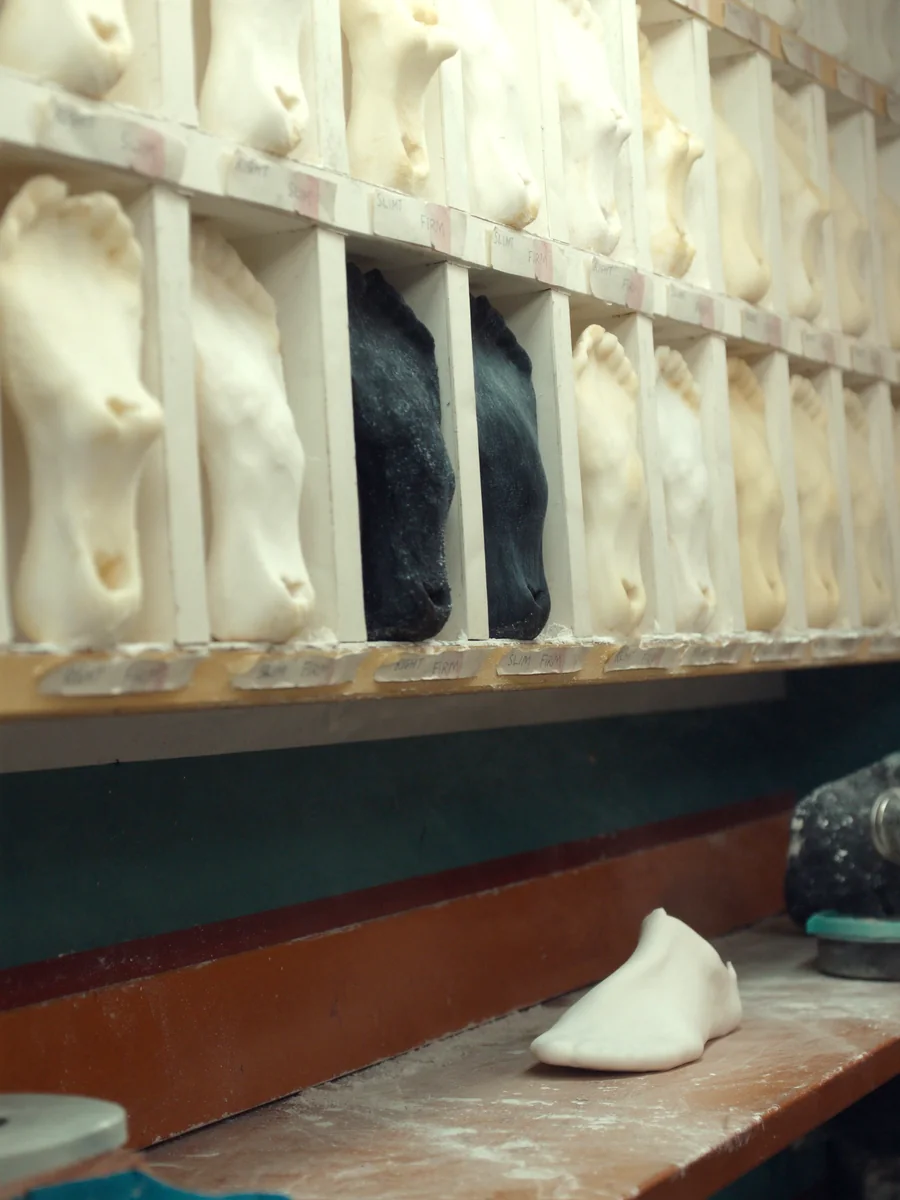 A wall rack of moulded orthotic shells in a clinic workroom, standing in labelled slots above a bench with a plaster foot cast.