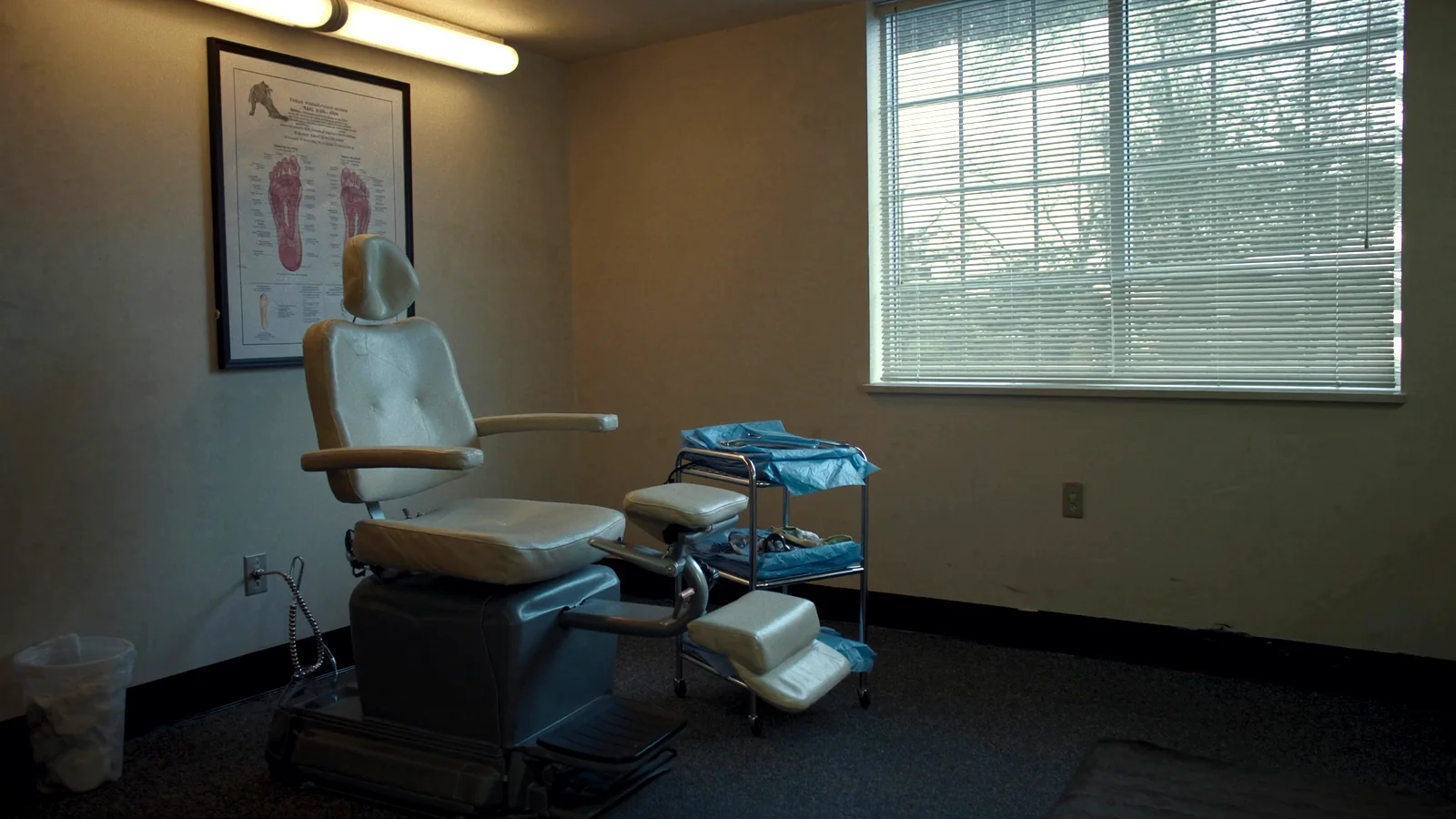 A podiatry treatment room with a hydraulic exam chair, its footrest extended, beside a rolling instrument tray under a window with half-open blinds.